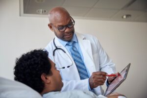 A male doctor in a white coat with a stethoscope around his neck, holding a tablet and explaining medical information to a patient lying in bed, inside a clinical setting.