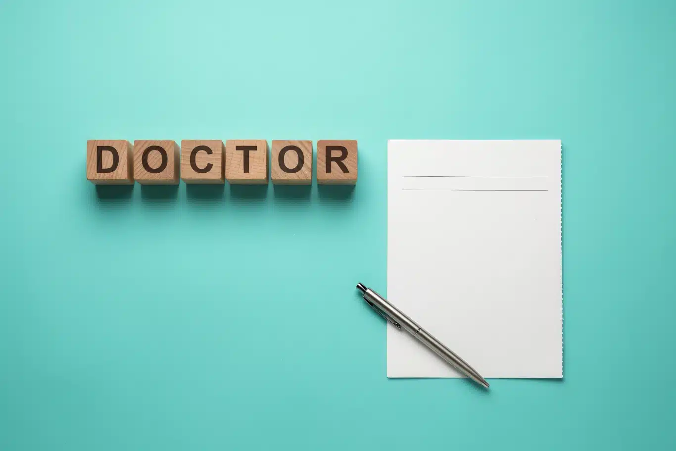 Wooden cubes spelling DOCTOR next to a blank notepad and pen Doctor spelled with blocks, next to a blank notepad and pen on a teal background